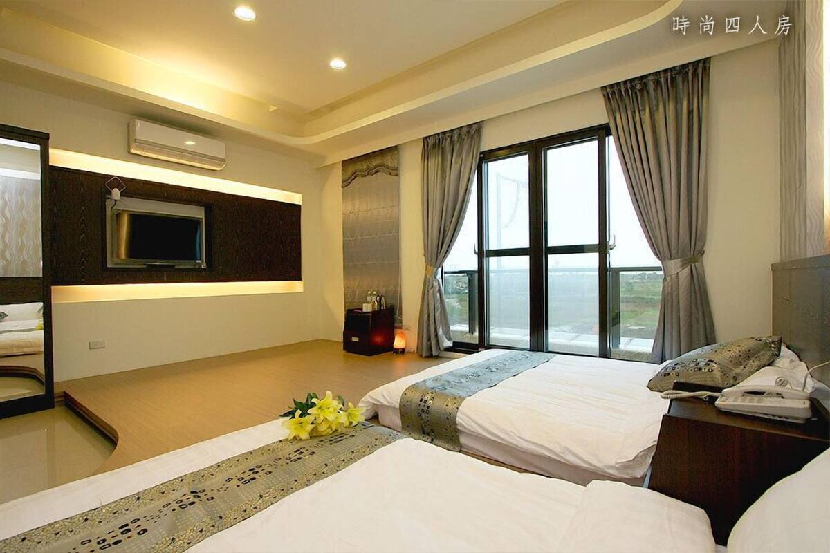 Habitación de hotel con dos camas, televisión, aire acondicionado, mesita de noche con lámpara y ventana con cortinas.