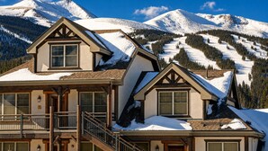 A chalet-style hotel with a balcony, wooden staircase, and a mountain backdrop.