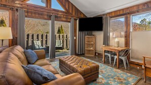 A spacious living room with a leather sofa, a wooden coffee table, and a dining area with a table and chairs. Large windows provide a view of the outdoors.