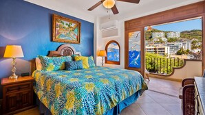 A bedroom with a bed, nightstands, a ceiling fan, a window with a view of buildings, and a balcony with wicker furniture.