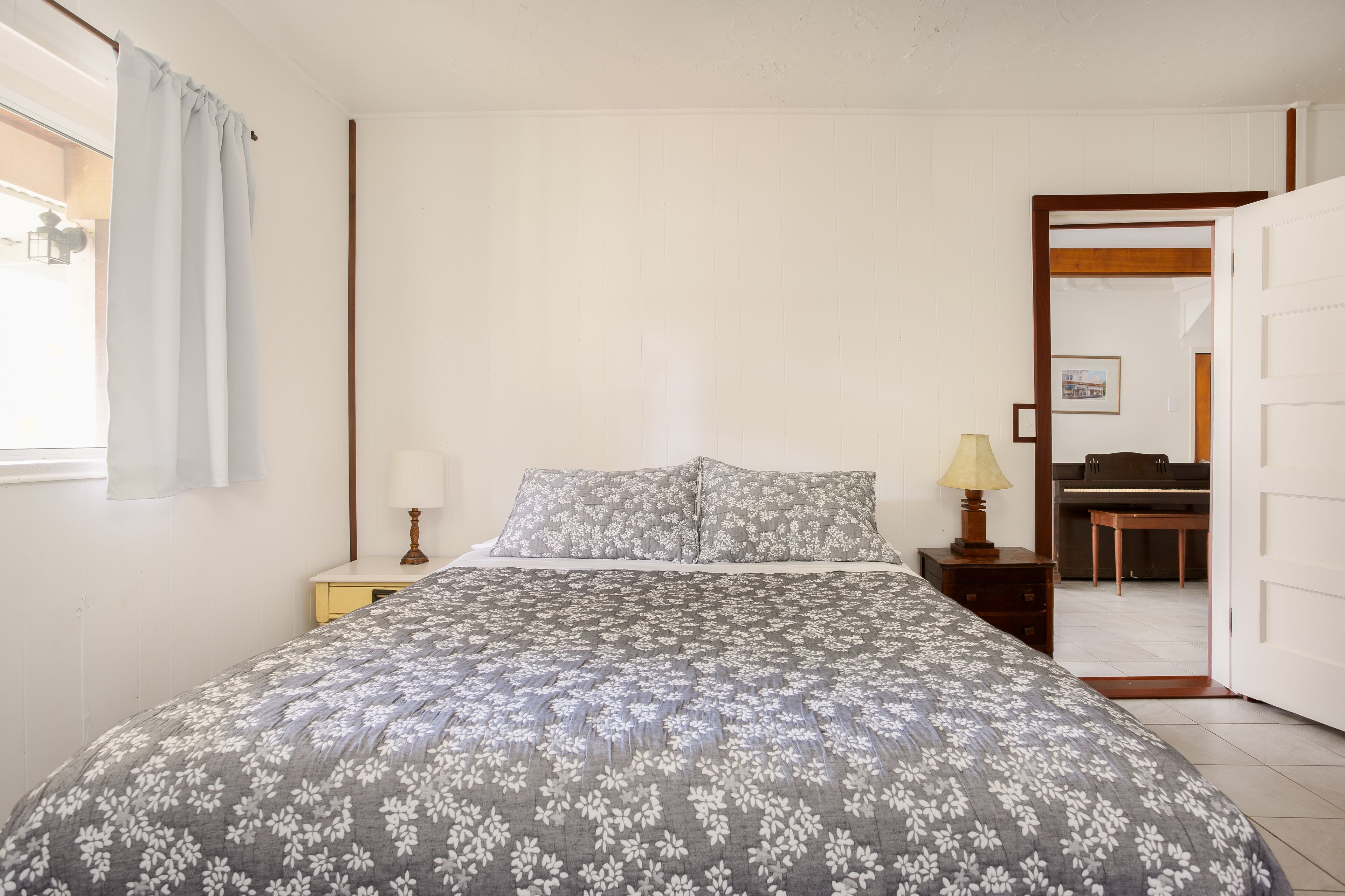 A bedroom with a bed, nightstand, lamp, and a view of the kitchen through an open door.
