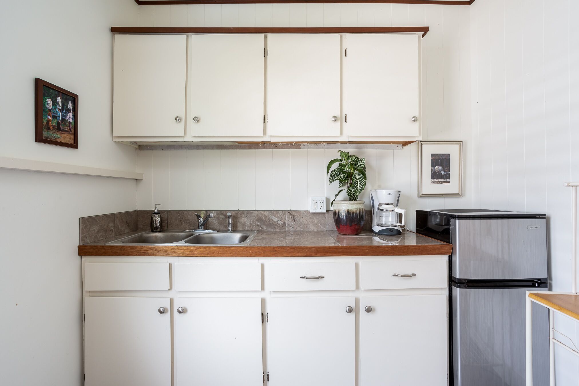 Una cocina con armarios blancos, un refrigerador de acero inoxidable, un lavamanos y una cafetera.