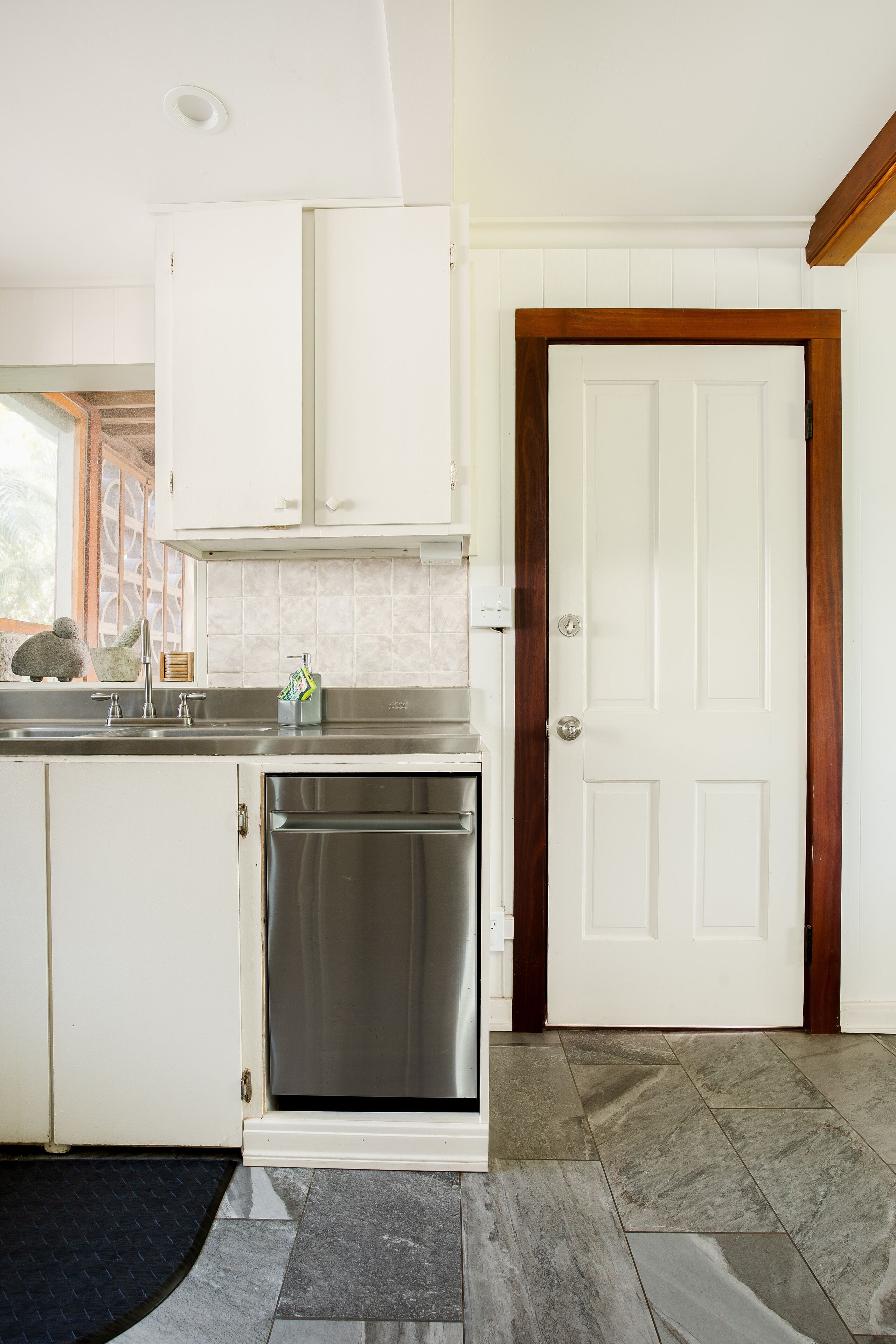 A kitchen with white cabinets, a stainless steel dishwasher, and a white door with a wooden frame.