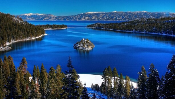 A large, clear lake surrounded by snow-covered mountains and dense forests.