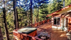 Een houten terras met een jacuzzi, zitplaatsen buiten, en een gebouw met een groot raam.