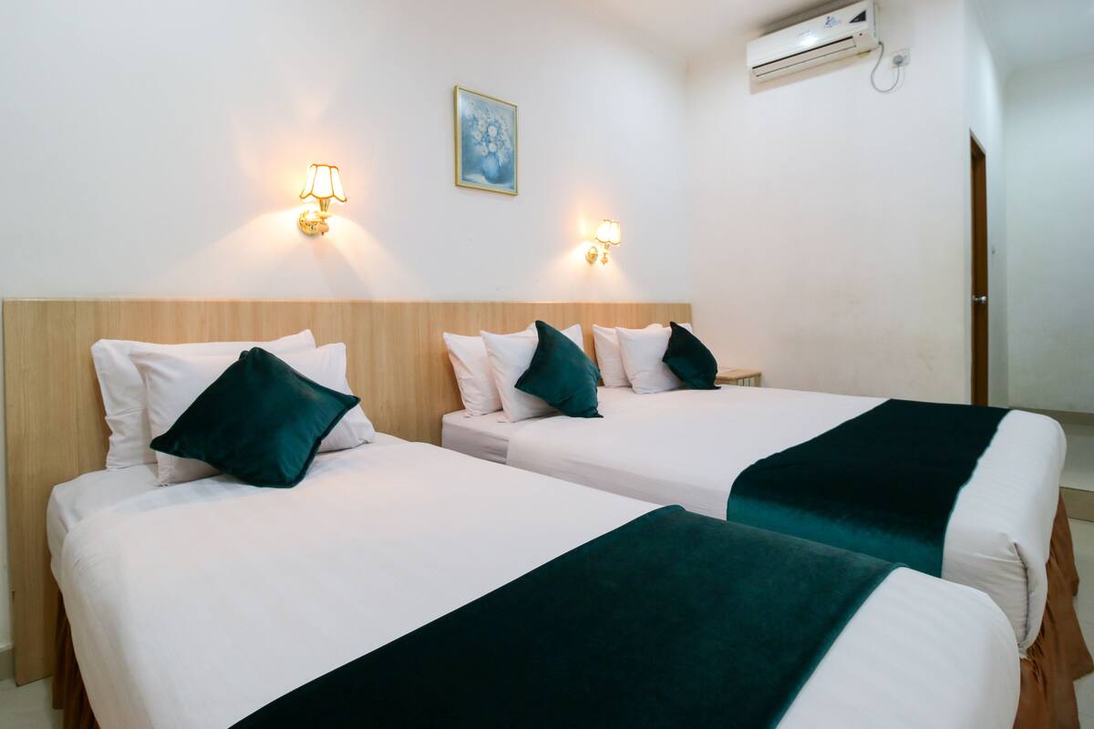 Ein Hotelzimmer mit zwei Einzelbetten, jeweils mit weißer Matratze, grünen Kissen und einer dunkelgrünen Decke. An der Wand sind Leuchten angebracht und über den Betten befindet sich eine Klimaanlage.