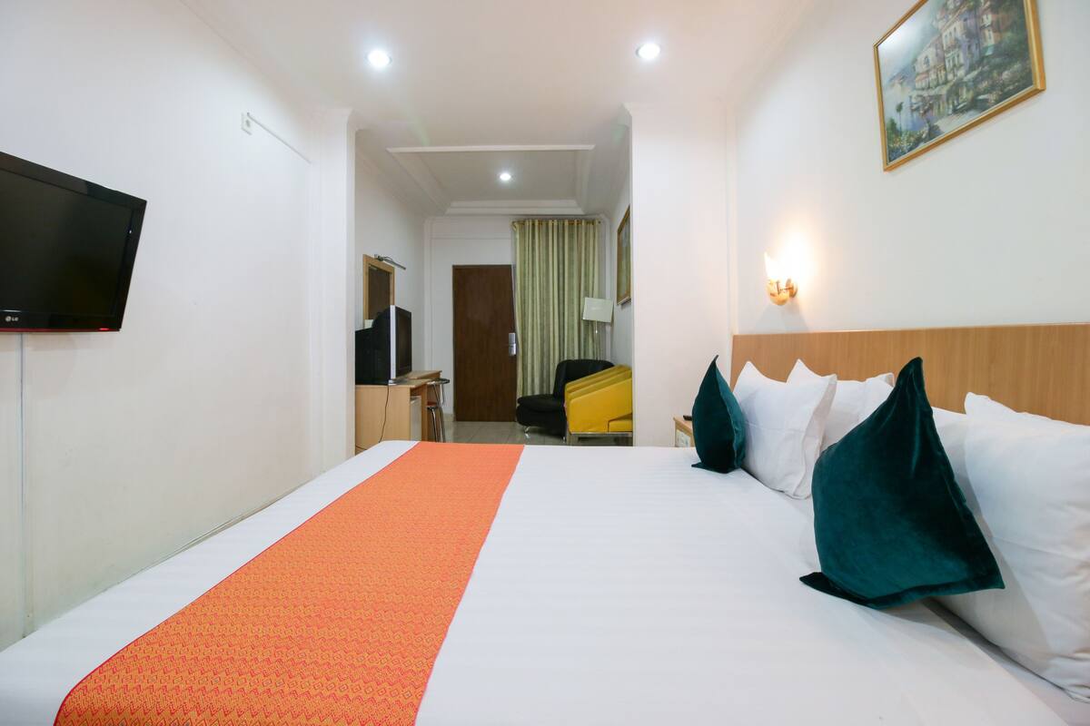 Ein Hotelzimmer mit einem großen Bett, einem flachbildfernseher an der Wand, einem Schreibtisch mit Stuhl und einer Tür zu einem weiteren Raum.