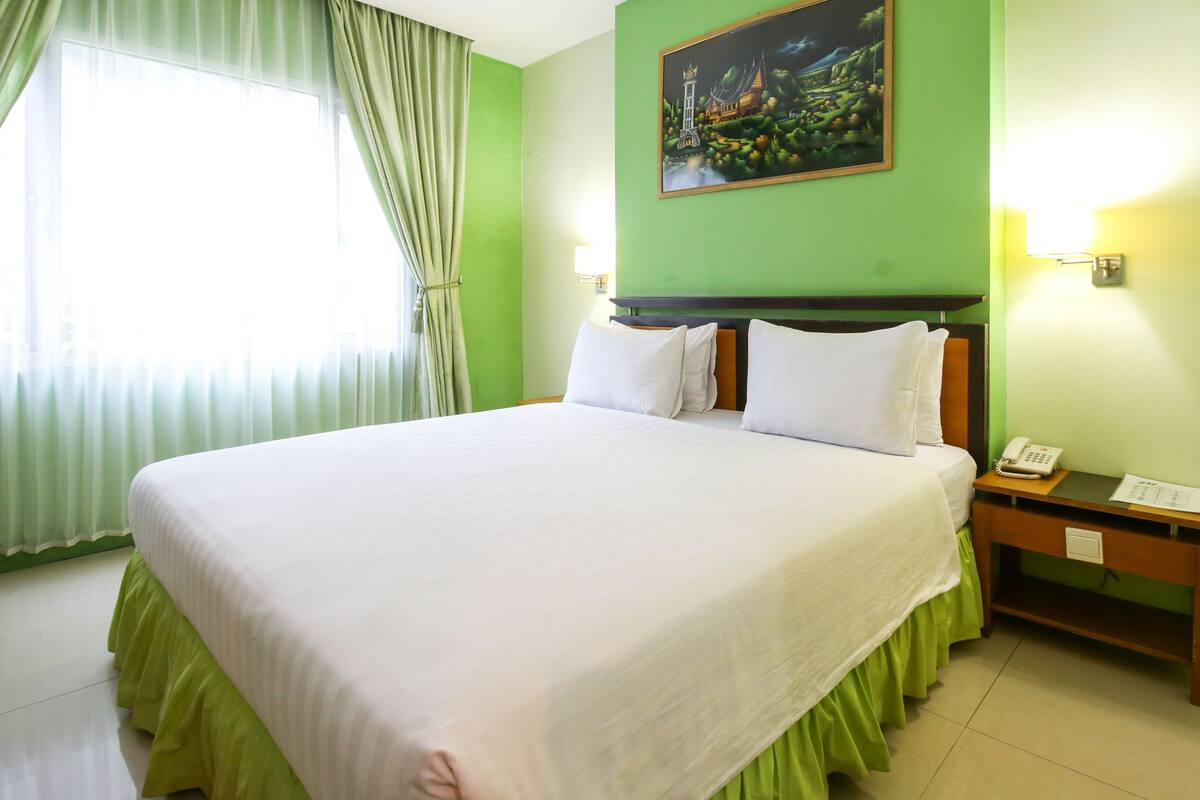 Ein Hotelzimmer mit einem großen Bett, grünen Vorhängen, einem Gemälde an der Wand und einem Nachttisch mit Telefon.