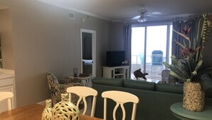 A living room with a green sofa, a wooden dining table with vases, a television on a stand, and a balcony with a view. - Unit #802 Beautiful Spacious Beachfront Condo! Sleeps 10! New Updates!! <br> (Orange Beach)