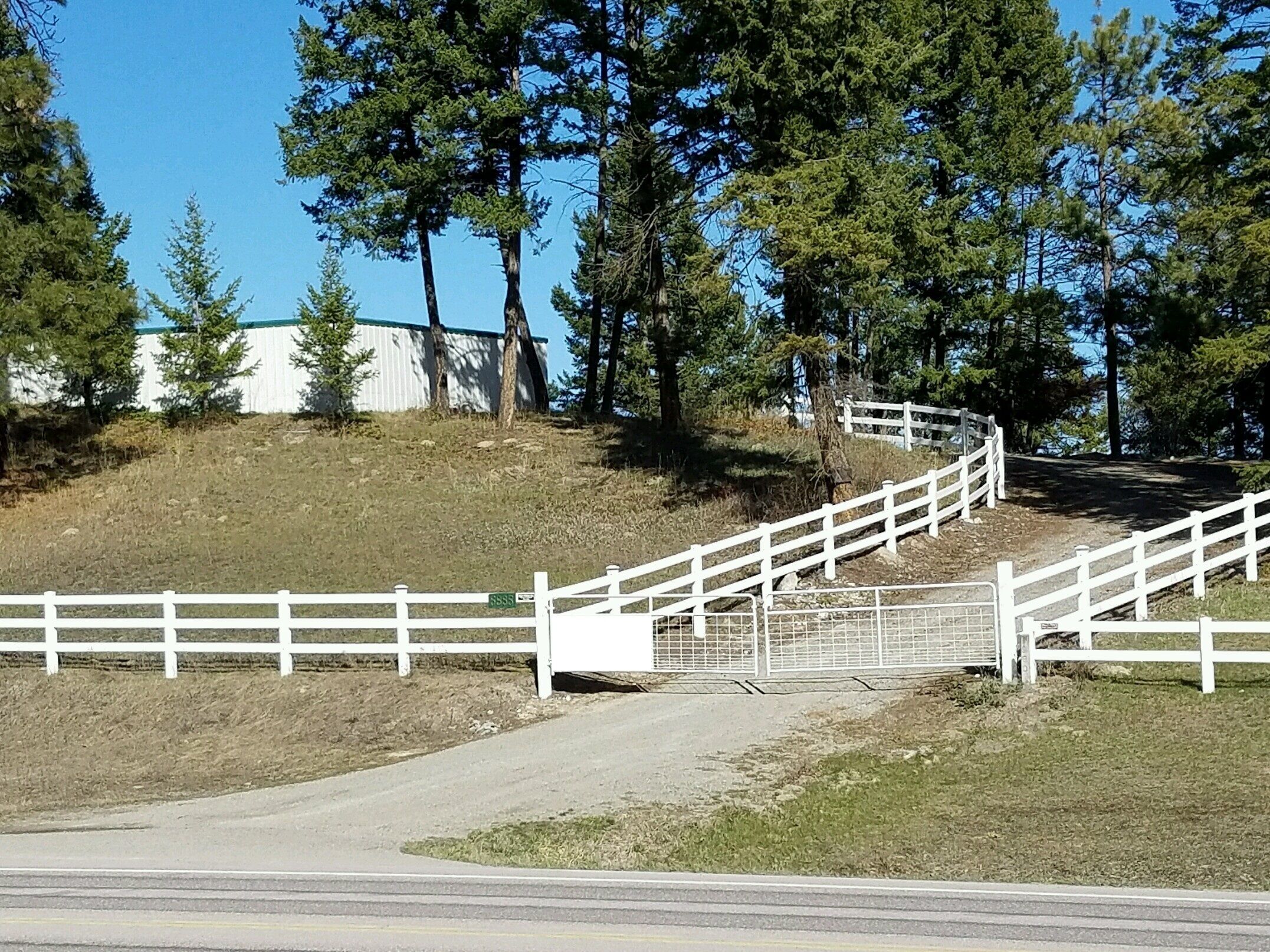 A white fence with a metal gate, a paved path, and a grassy hill with trees.
