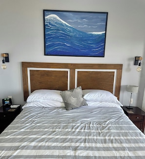 Una camera d'albergo con un letto grande, una testiera in legno, un comodino con una lampada e un quadro appeso al muro raffigurante un'onda.