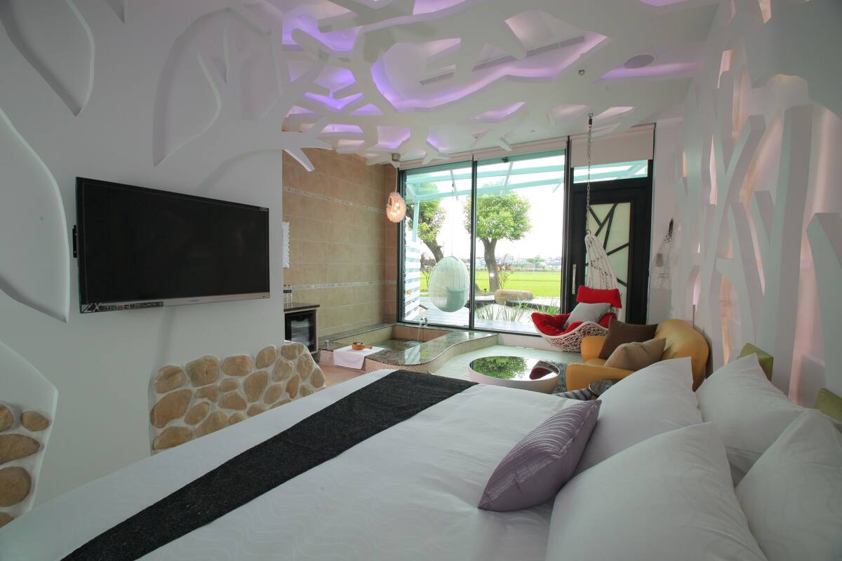 Quarto de hotel moderno com uma cama grande, televisão de tela plana, área de estar com uma poltrona vermelha e vista para o exterior.