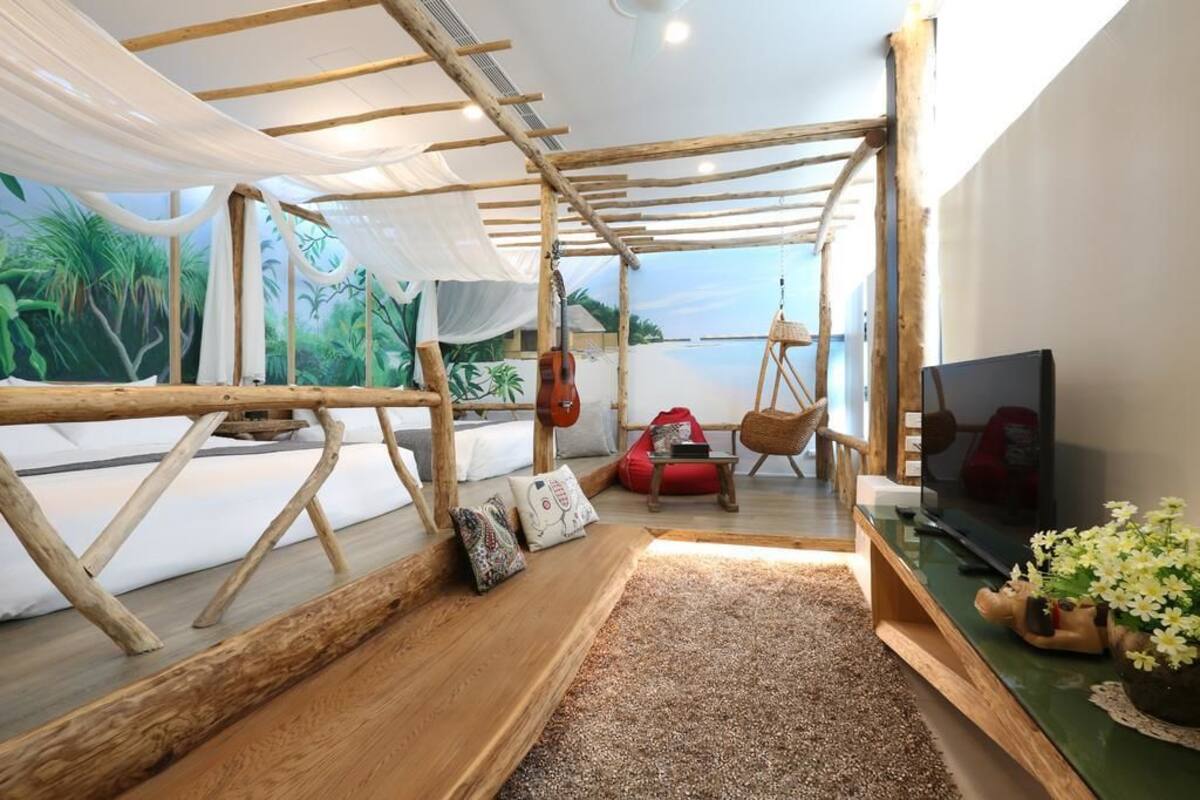 Quarto com estrutura de teto em madeira, cama com roupa de cama branca, televisão sobre um suporte de madeira e vista para o verde pela janela.