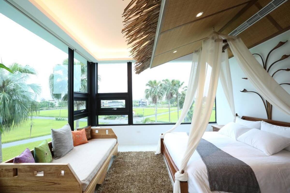 Quarto com uma janela grande, uma cama com roupa de cama branca, um banco de madeira com almofadas e vista para uma paisagem verde.