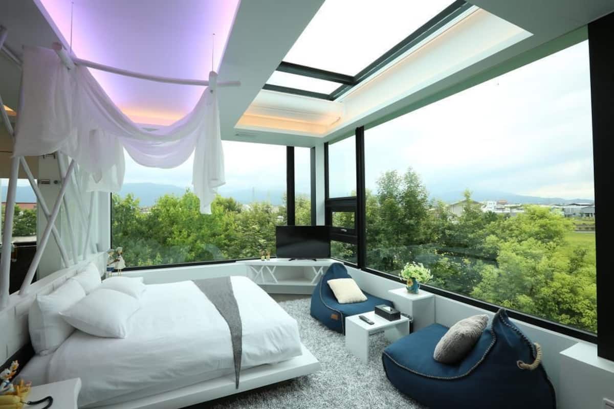 Quarto moderno com uma cama grande, área de estar e televisão. Possui uma clarabóia e vista para o verde.