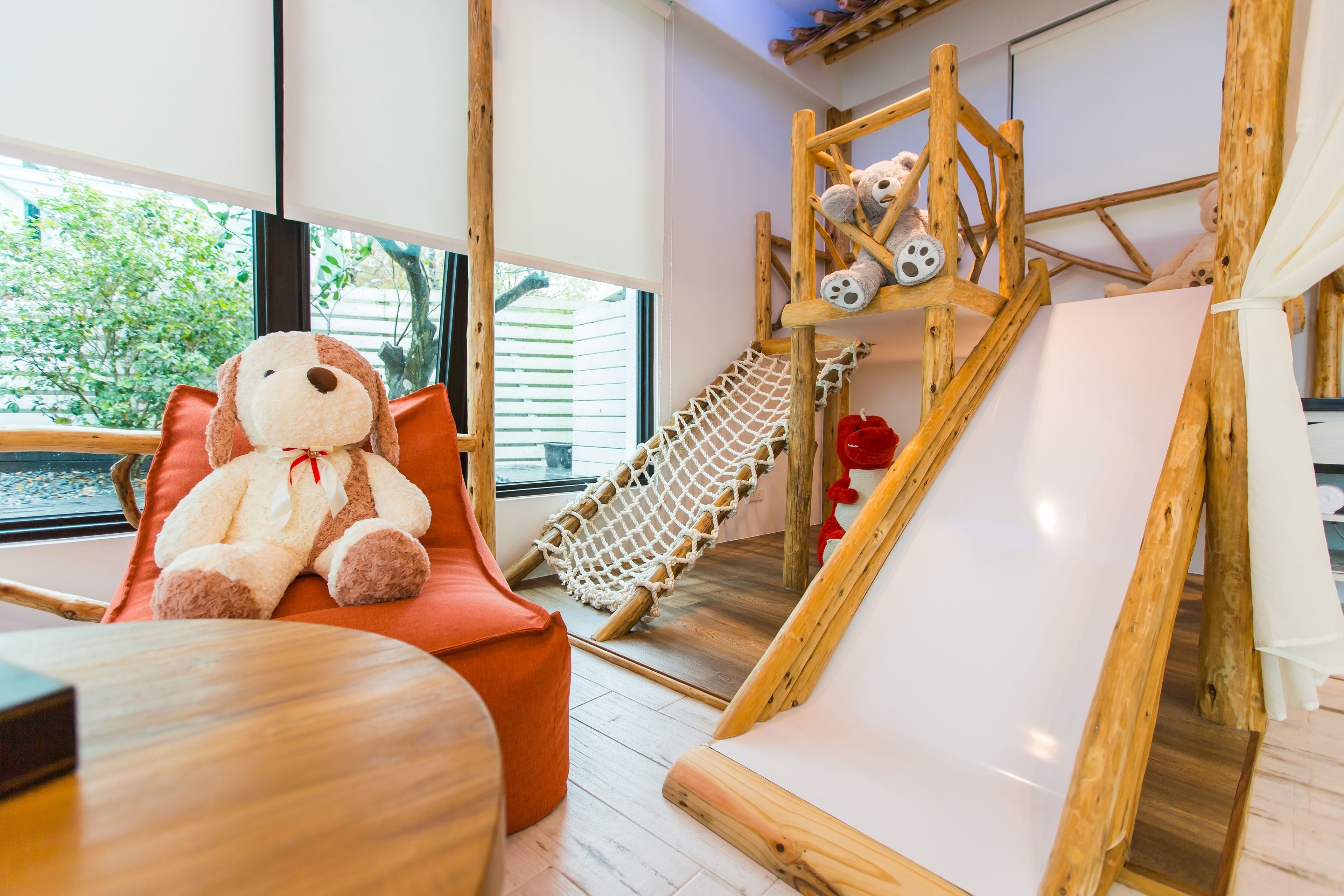 Una habitación acogedora con un gran oso de peluche, una hamaca, un parque infantil de madera y una pizarra.