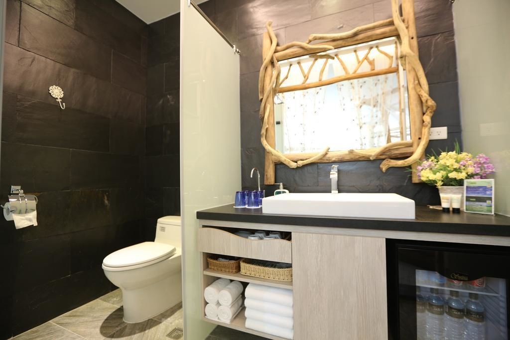 Un baño con pared de azulejos oscuros, un inodoro blanco, un lavabo con espejo de marco de madera y una estantería con toallas y artículos de aseo.
