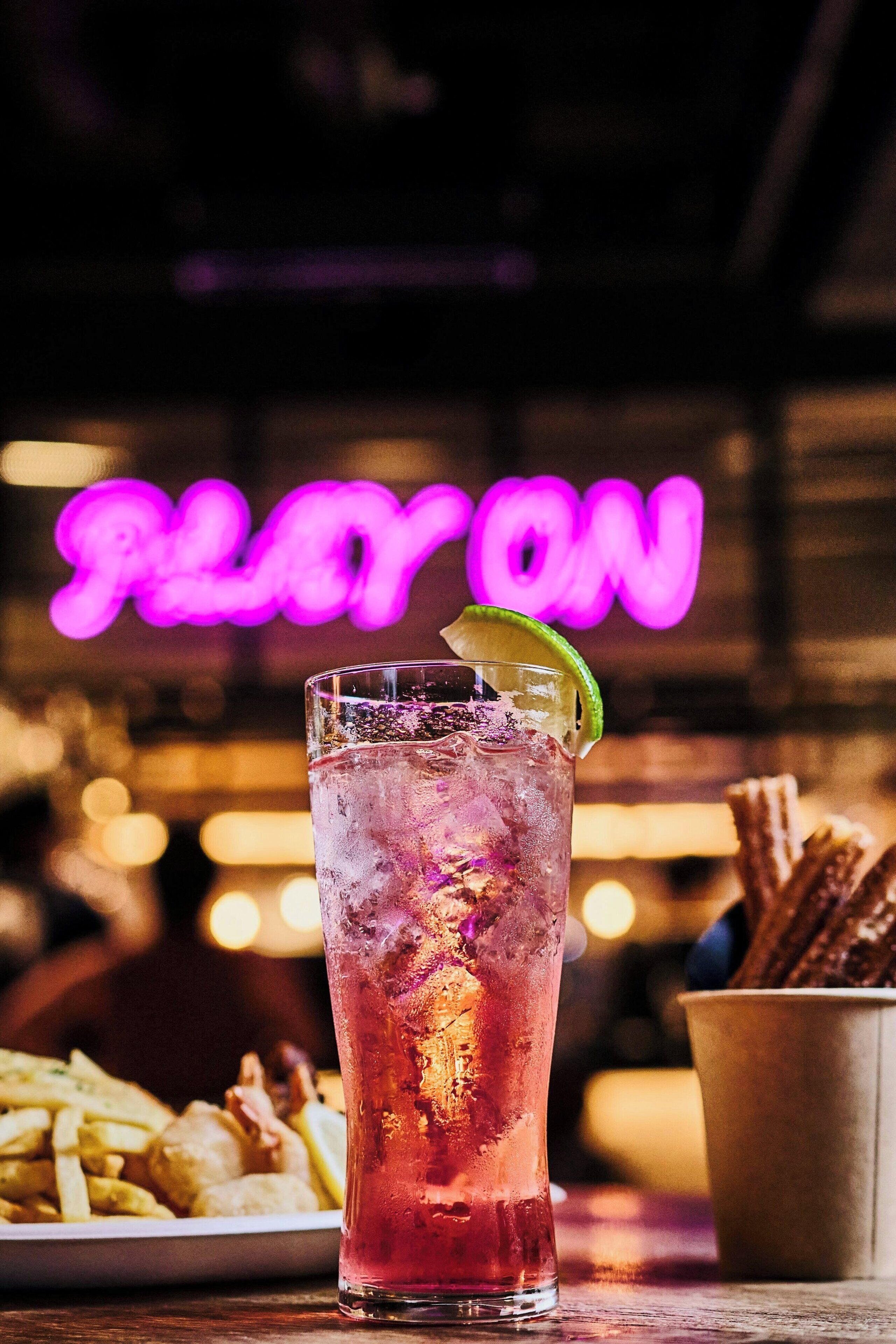 A tall glass with a lime wedge, ice cubes, and a pink beverage, possibly a cocktail, on a wooden table.