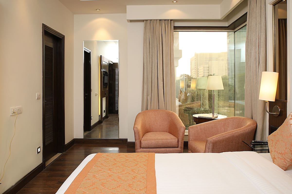 Quarto de hotel com uma cama grande, duas poltronas, uma pequena mesa com abajur, e vista da cidade através de uma janela ampla.