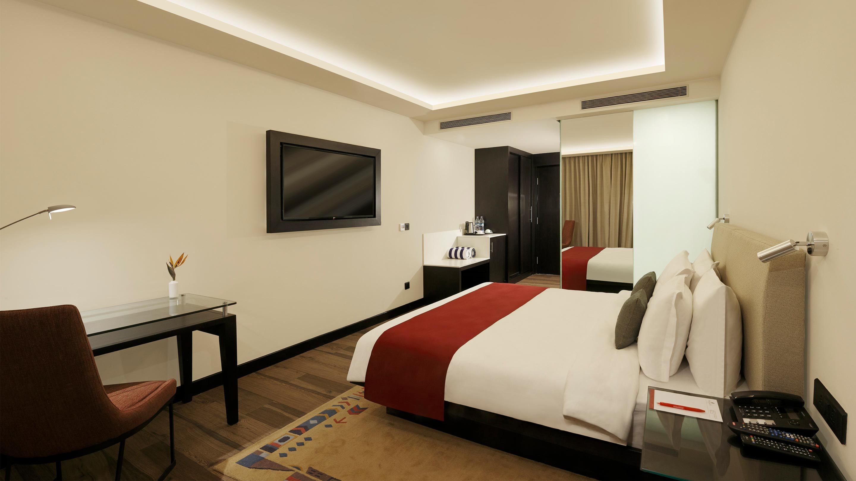 A hotel room with a bed, a desk, a chair, a TV, a mirror, and a telephone.