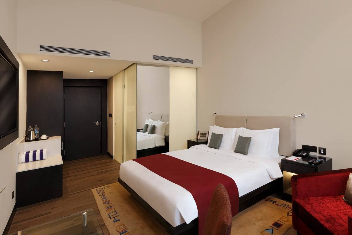 A hotel room with a large bed, a TV mounted on the wall, a bedside table with a lamp, and a red armchair.
