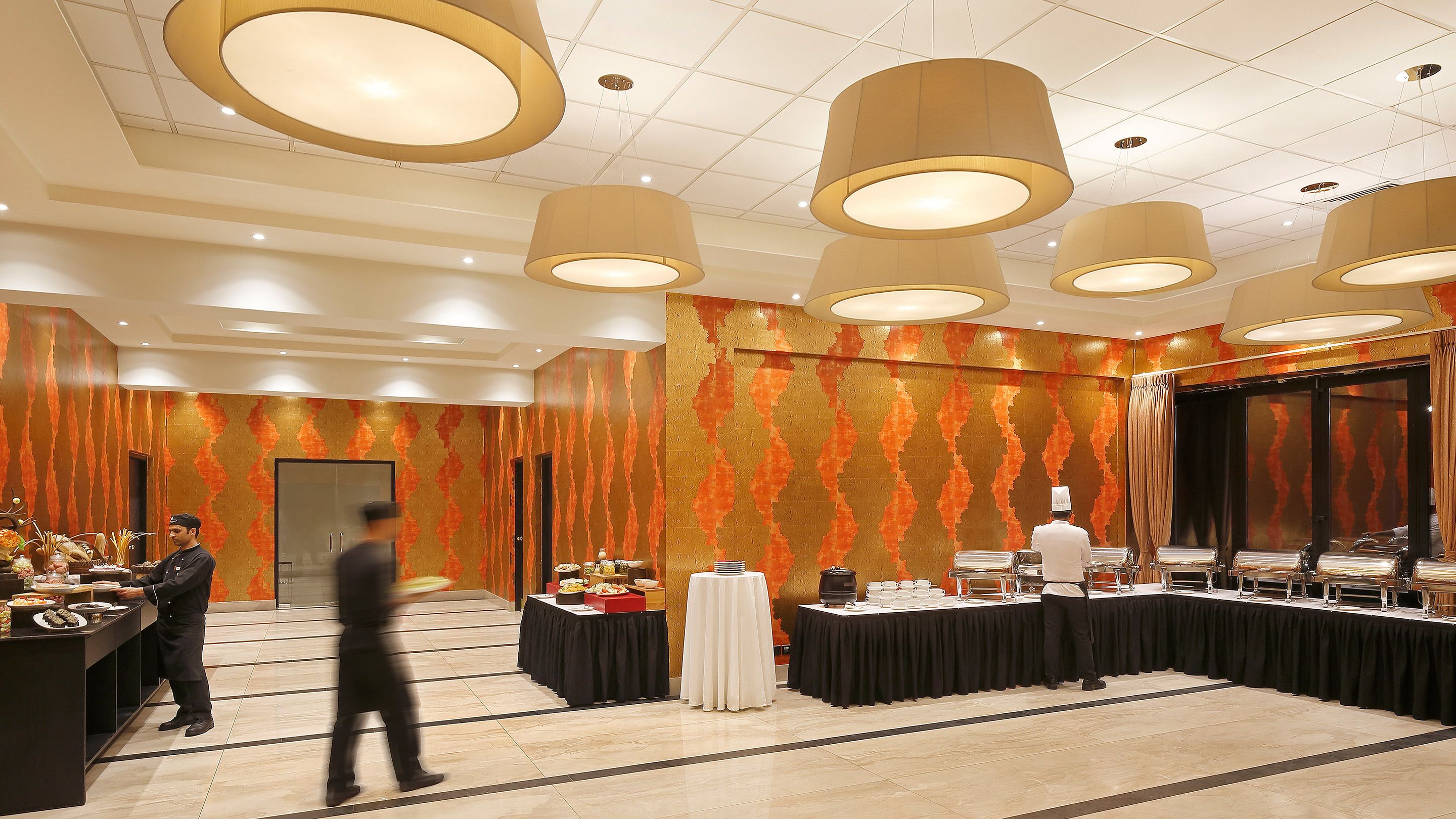 Une salle de banquet avec une longue table dressée pour un repas, un espace buffet où sont présentés différents plats, et une scène avec des rideaux.
