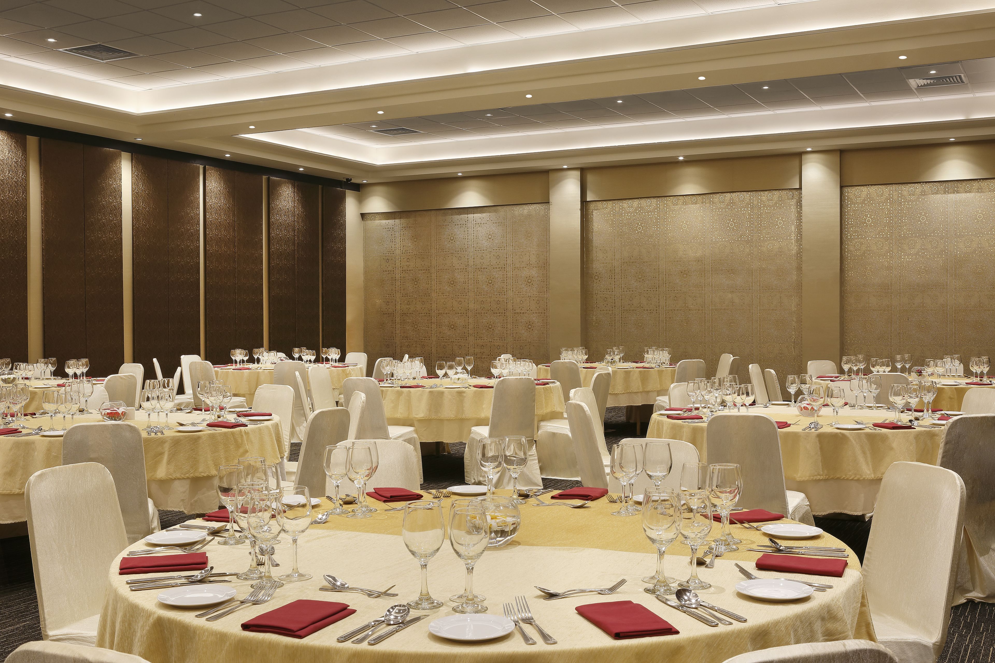 A banquet hall with round tables set for dining, equipped with cutlery, glasses, and red napkins.