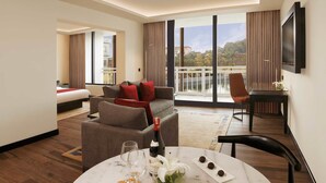 Habitación de hotel con cama, sofá, mesa de comedor con una botella y vasos, y vista a un balcón con barandilla.