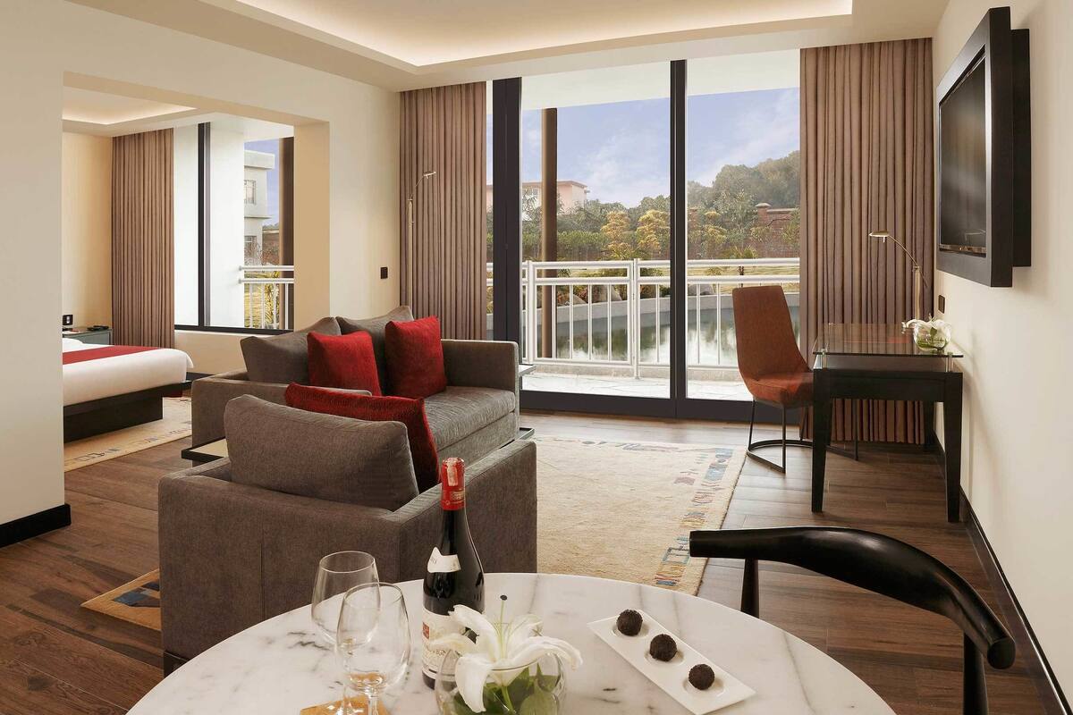 A hotel room with a bed, a sofa, a dining table with a bottle and glasses, and a view of a balcony with a railing.