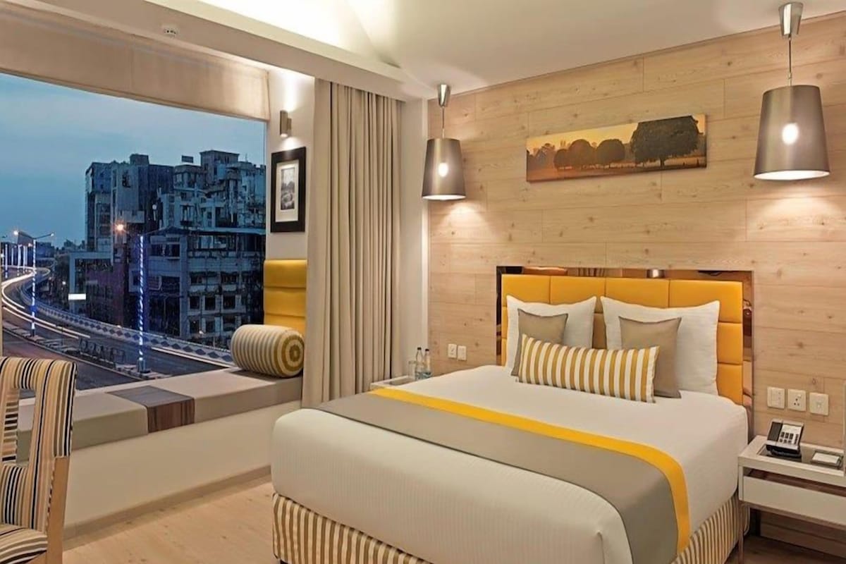 Una habitación de hotel moderna con una cama grande, vistas a la ciudad, un cabecero de madera y una mesita de noche con un teléfono.