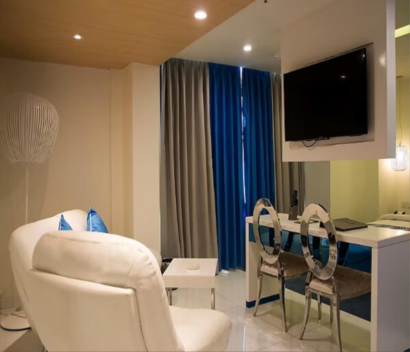 Una sala de estar moderna con un sofá blanco, una mesa de comedor con sillas, un televisor de pantalla plana montado en la pared y un amplio ventanal con cortinas azules.