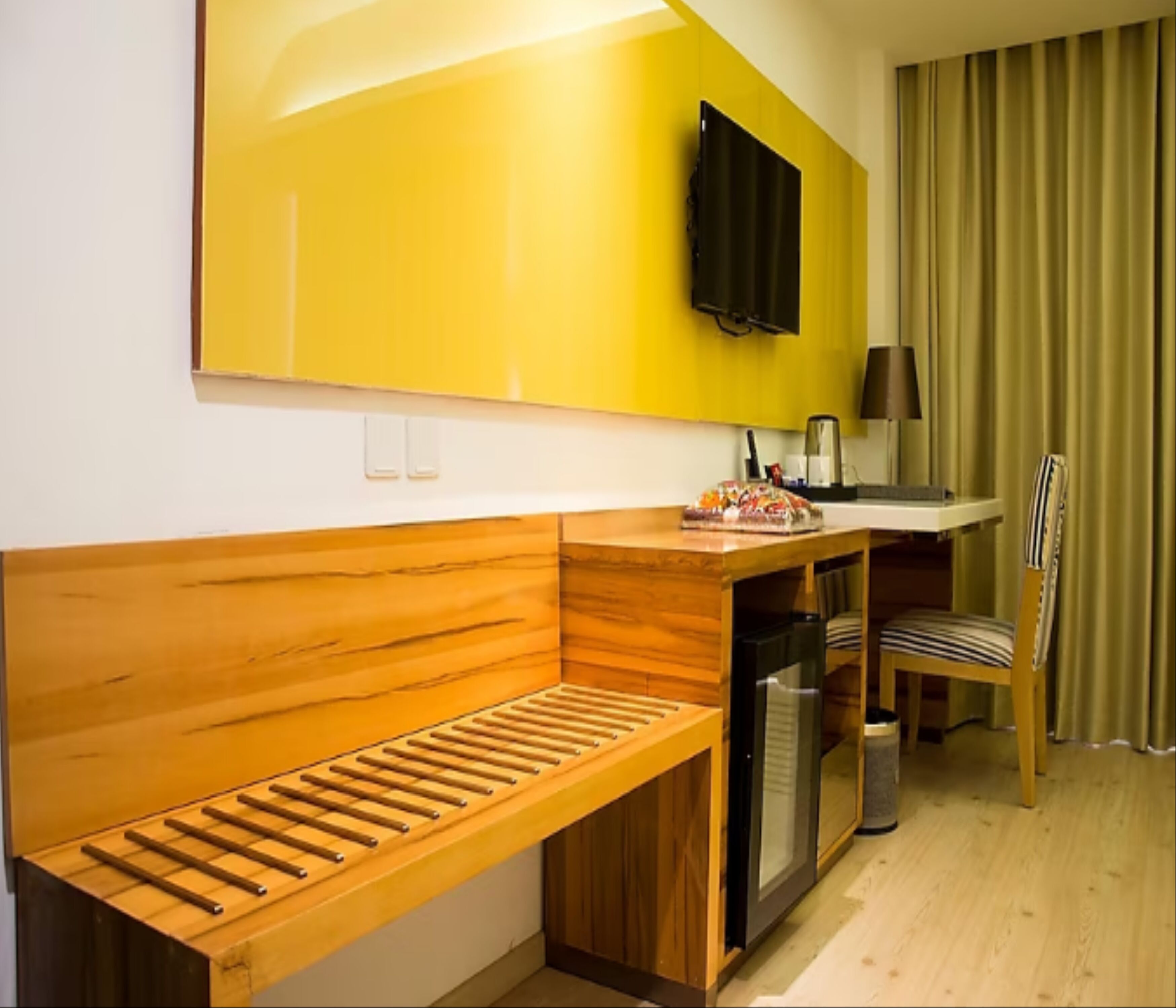 A compact hotel room with a wooden bench, a small kitchenette, a television, and a window with curtains.