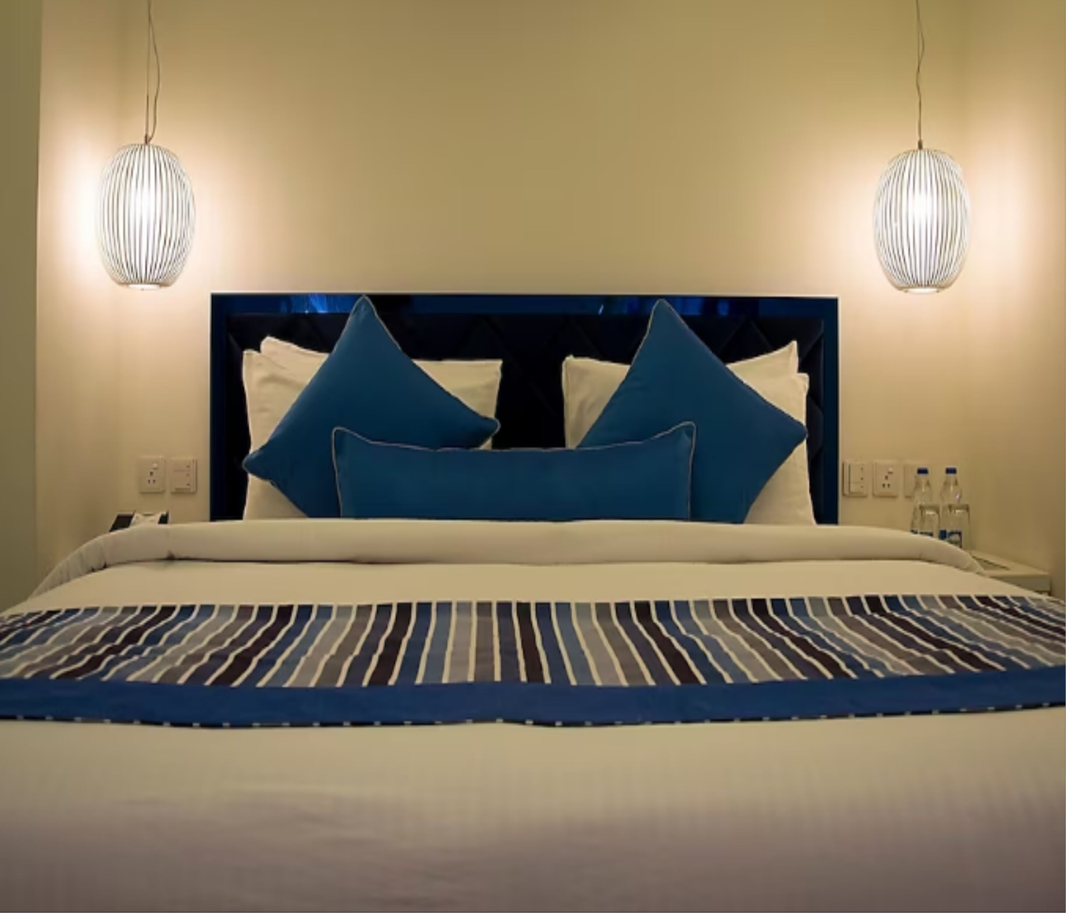 Cama bem arrumada com colcha listrada e almofadas azuis, duas luzes pendentes e uma tomada na parede.
