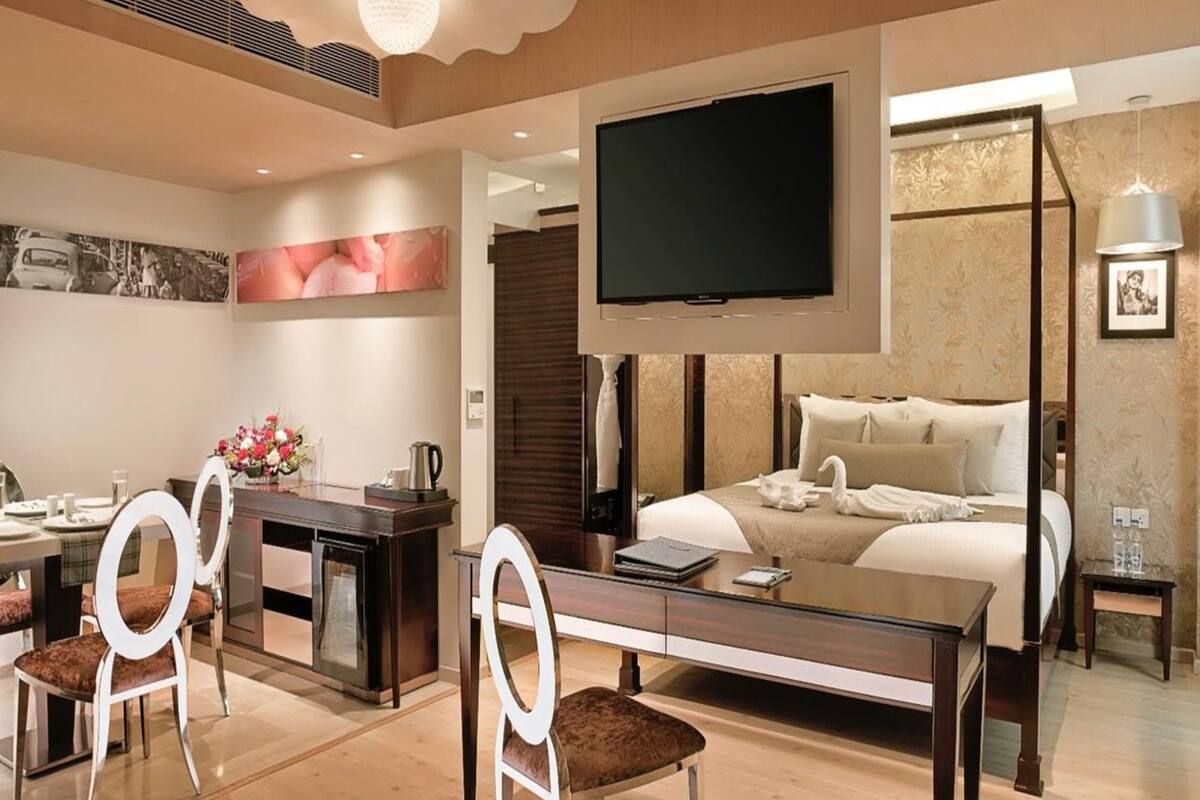 A modern hotel room with a large bed, a flat-screen TV, a dining area with chairs and a table, and a decorative ceiling light.