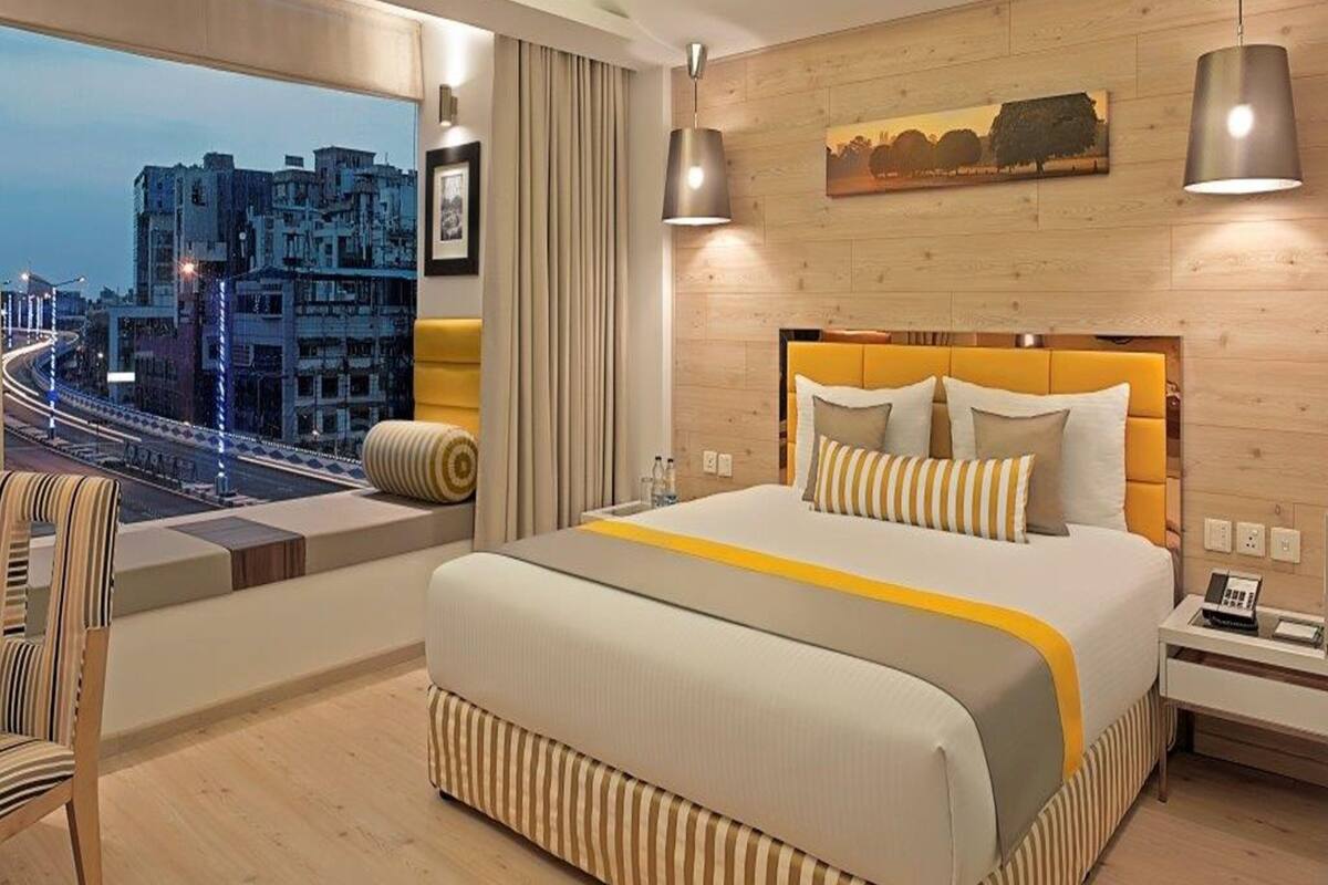 A modern hotel room with a large bed, a view of the cityscape, a wooden headboard, and a nightstand with a phone.