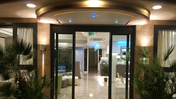 A hotel entrance with a modern design, featuring a glass door, a reception desk, and a seating area with chairs and a table. - Hotel Adele (Bellaria-Igea Marina)