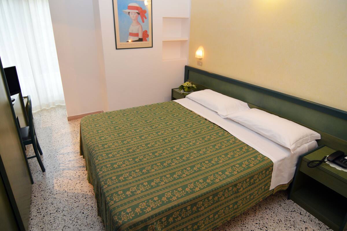 Una habitación de hotel con una cama, una silla, una mesita de noche con un teléfono y un cuadro en la pared.