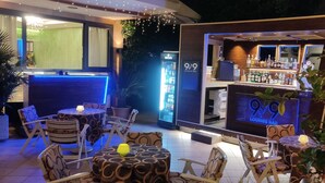 An outdoor lounge area with tables and chairs, a bar with a neon sign, and a refrigerator. - Hotel Adele (Bellaria-Igea Marina)