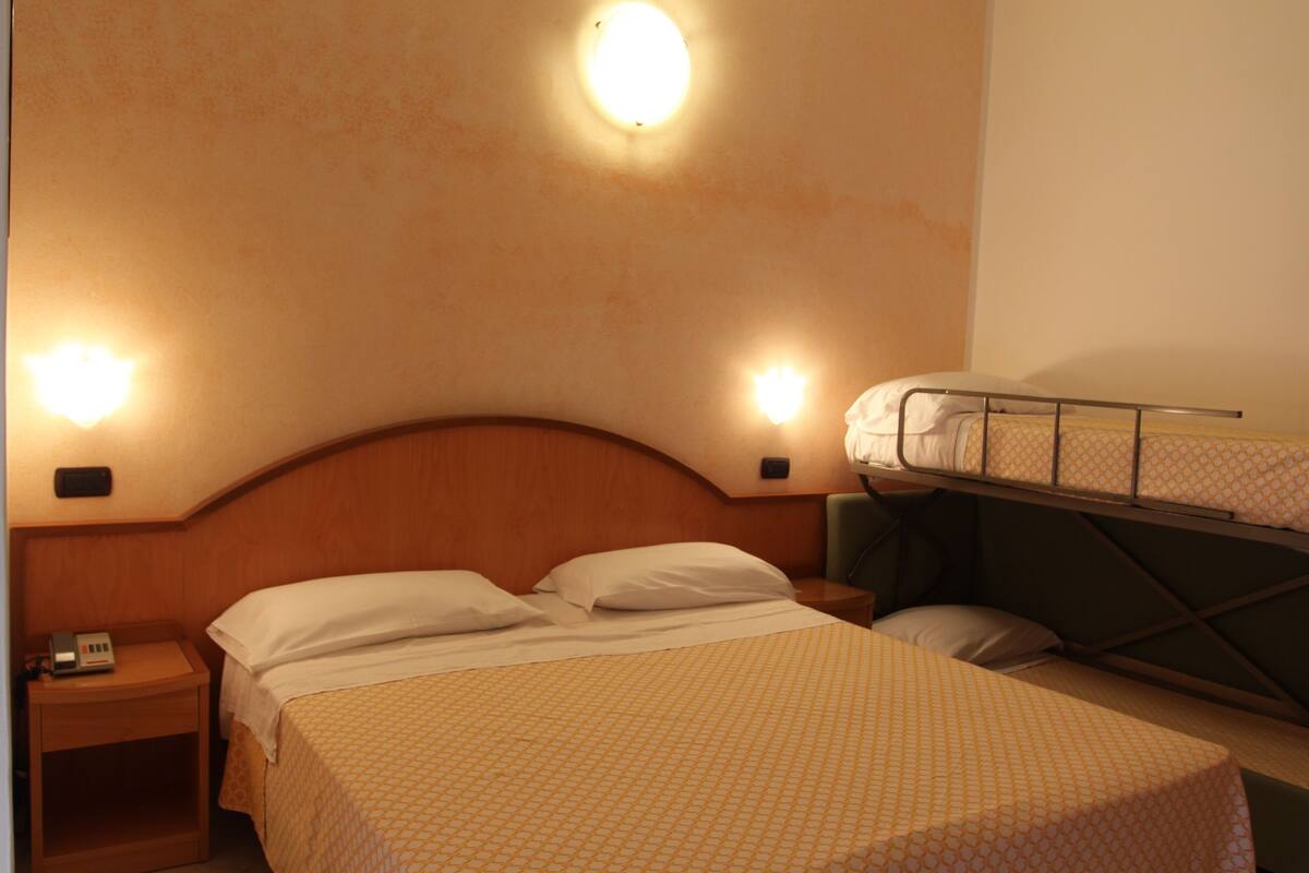 Habitación de hotel con una cama individual, una litera, una mesita de noche con un teléfono y dos luces fijadas a la pared.