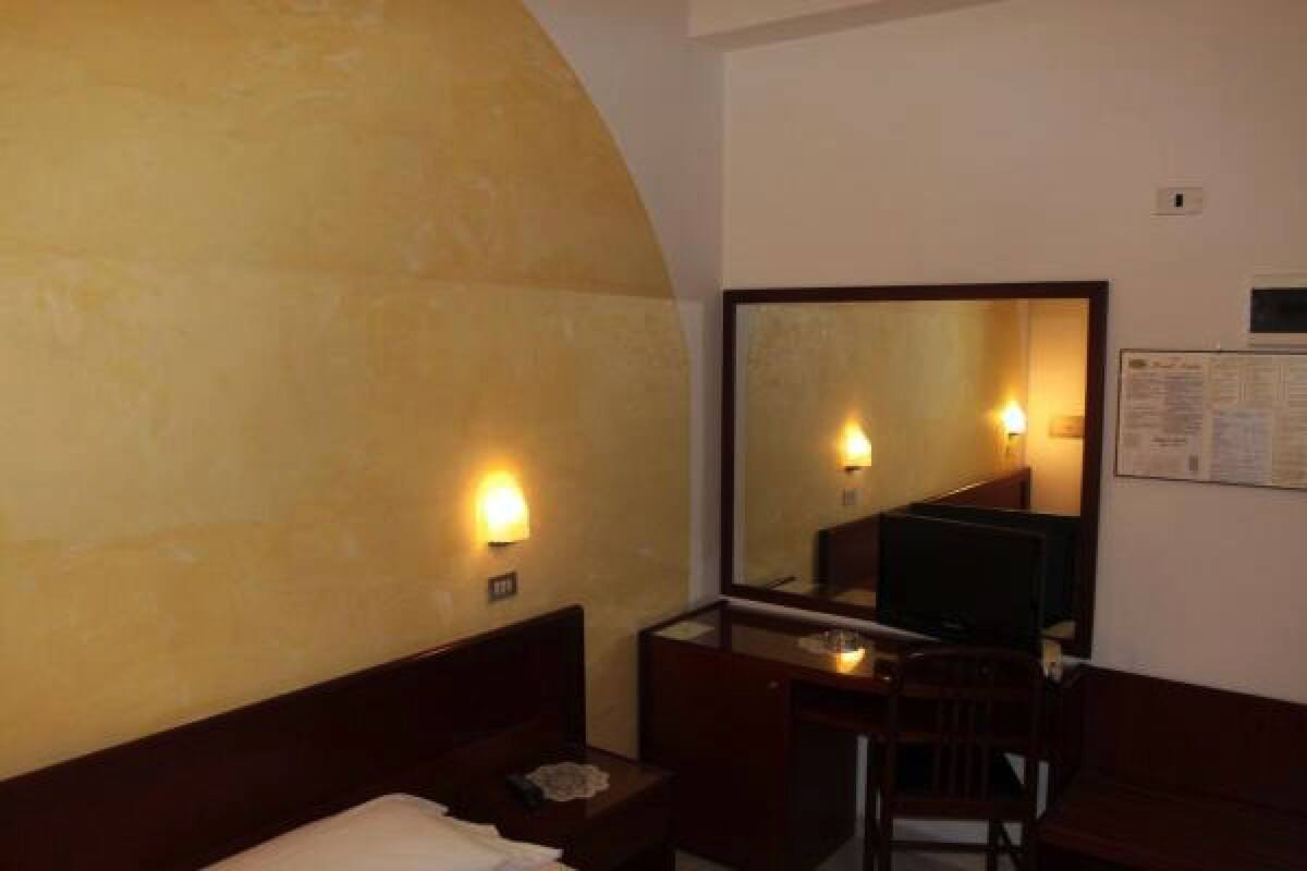 Una habitación de hotel con cama, escritorio, silla, espejo y un tablón de anuncios en la pared.