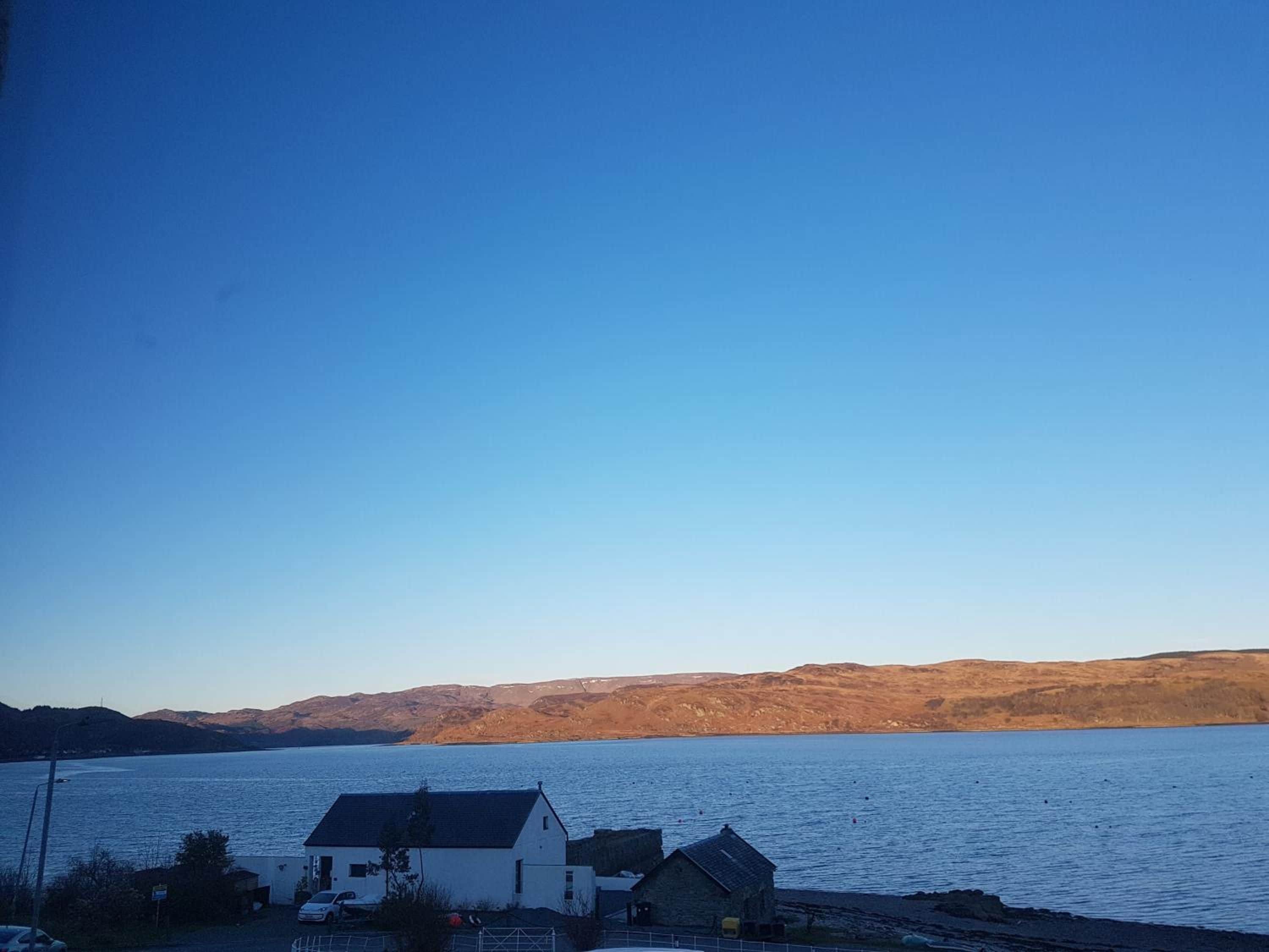 Vista costera con un edificio blanco y una estructura más pequeña, posiblemente un cobertizo, cerca de una masa de agua con colinas al fondo.