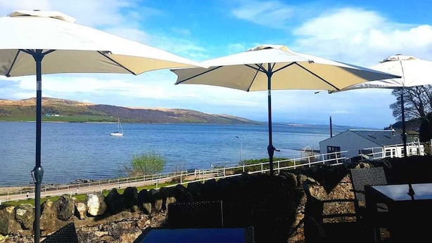 Een terras aan de kust met buitenmeubels en parasols met uitzicht op een waterpartij.