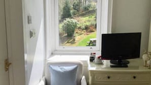 Een kleine, goed verlichte kamer met een enkele witte fauteuil, een televisie op een witte dressoir en een raam met uitzicht op een tuin.