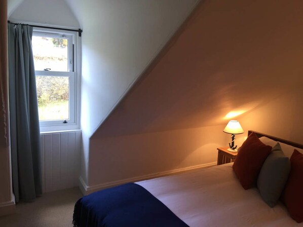 A bedroom with a bed, a nightstand with a lamp, a window with curtains, and a view of the outdoors. - Kames Hotel (Tighnabruaich)