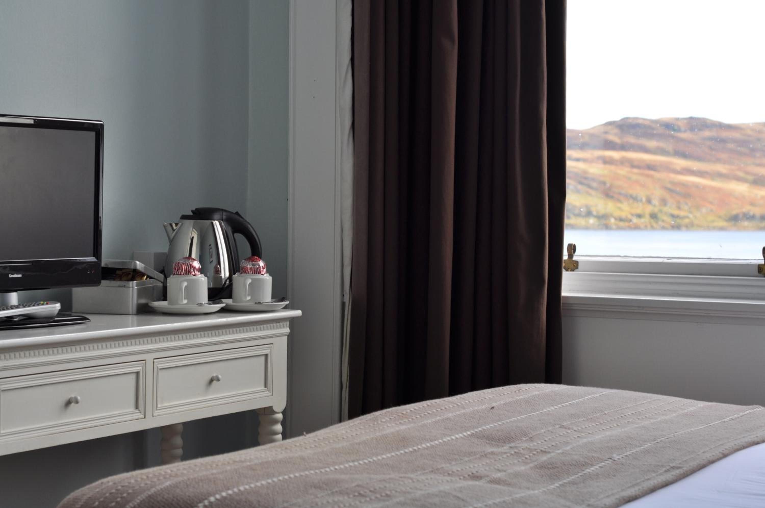 Una habitación de hotel con cama, escritorio con televisor, cafetera y vista a un lago y colinas.