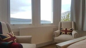 Een kamer met twee fauteuils en een bed, grote ramen met uitzicht op zee en een radiator onder de ramen.