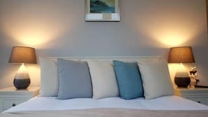 Een net opgemaakt bed met een witte dekbedovertrek en vier kussens in blauw- en beigetinten. Aan beide zijden van het bed staan nachtlampjes, en boven het hoofdeinde hangt een ingelijst schilderij.