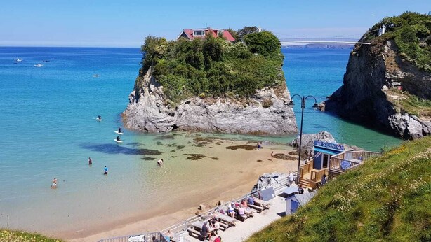 Une plage côtière aux eaux bleues et limpides, un promontoire rocheux sur lequel se trouve une maison, et une jetée où se trouvent des personnes.