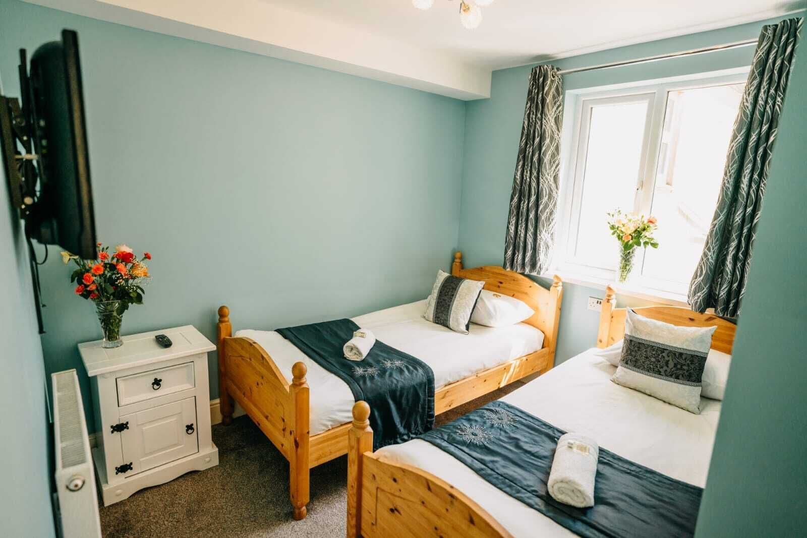 A bedroom with two wooden beds, a white bedside table, a TV mounted on the wall, and a window with curtains.
