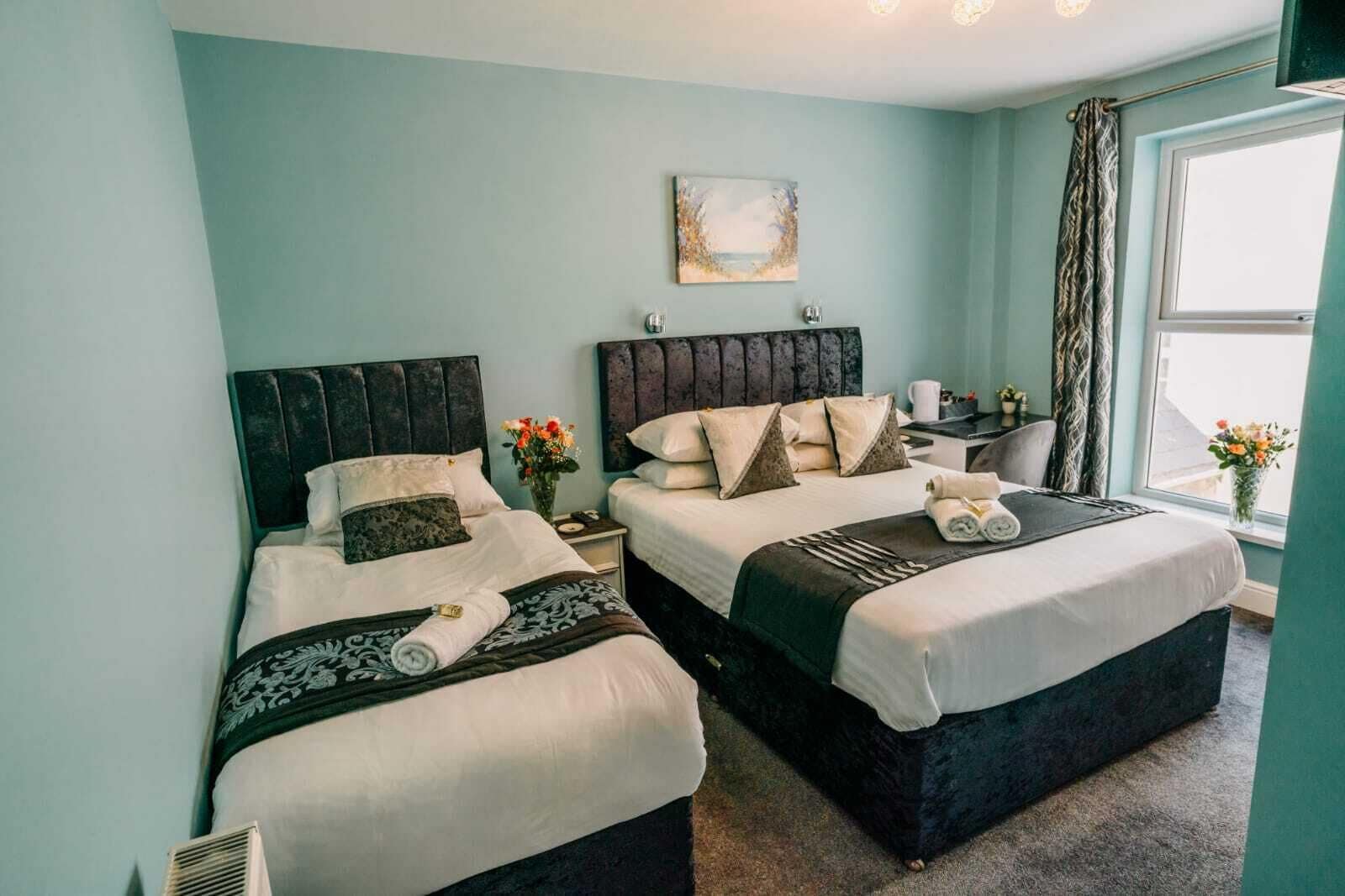 A hotel room with two beds, a window with curtains, a bedside table with a lamp, and a painting on the wall.